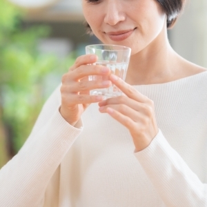 Close-up of a smiling woman wearing a light sweater, holding a glass of water with both hands.