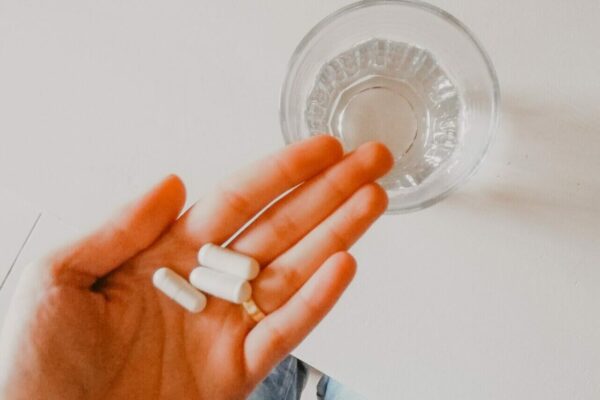 Hand holding three white capsule pills over a glass of water on a white table surface